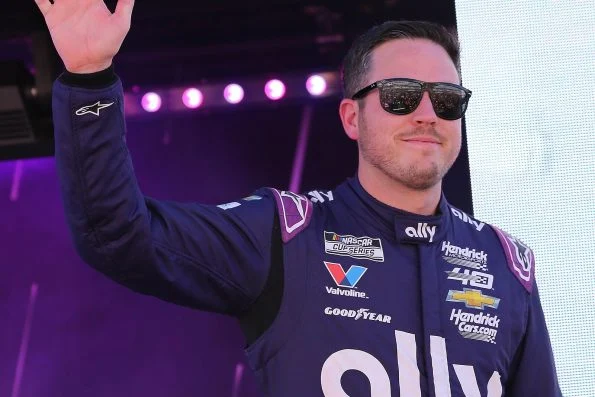 Alex Bowman in a purple Ally race suit and sunglasses, raising a hand during driver introductions under stage lighting.