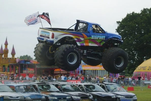 Bob Chandler creator and founder of Bigfoot monster truck airborne above a long row of scrap cars at an outdoor show.