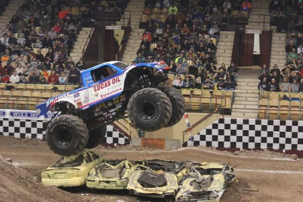 Bob Chandler's blue Bigfoot 15 monster truck mid-jump over crushed cars in a packed arena.