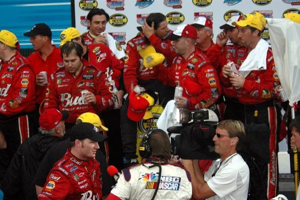 Dale Earnhardt Jr. being interviewed in victory lane, surrounded by his Budweiser crew in red uniforms, with a trophy alongside.