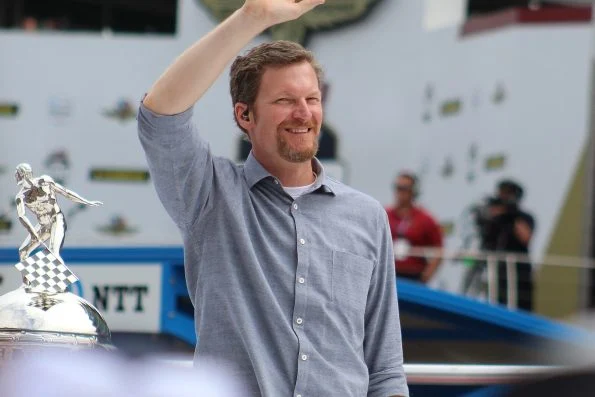 Dale Earnhardt Jr. in a grey shirt raising a hand to wave, with photographers and a trophy visible behind him.