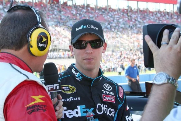 Denny Hamlin in a FedEx cap and race suit being interviewed by a television reporter at Auto Club Speedway, grandstands behind.
