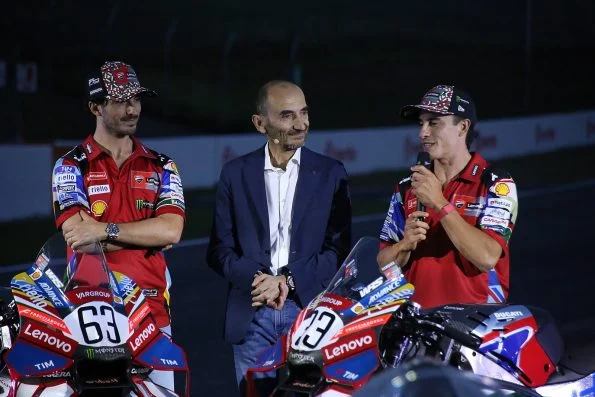 Francesco Bagnaia on the left with his arms folded and Marc Márquez on the right holding a microphone, either side of Ducati chief executive Claudio Domenicali, behind their number 63 and 93 machines.