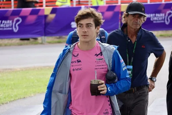 Franco Colapinto in a pink Alpine team shirt and blue jacket walking into the circuit, carrying a drink.