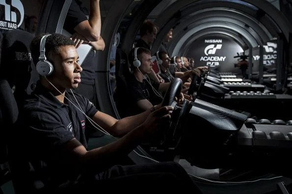 Jann Mardenborough seated at a Nissan GT Academy racing simulator wearing a headset, gripping the wheel, with other competitors at rigs behind him.
