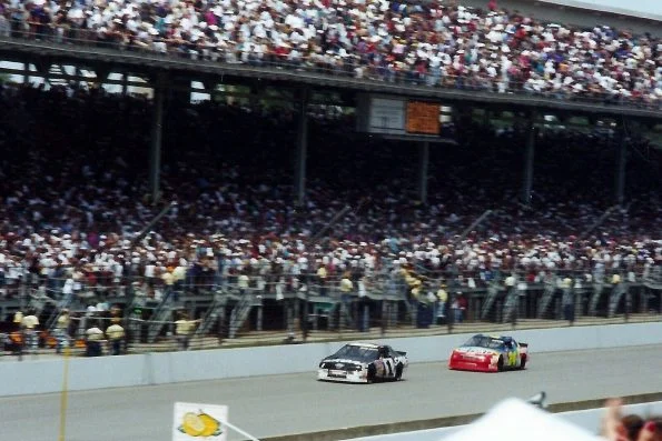 Jeff Gordon's number 24 car following Rick Mast's number 1 on track at Indianapolis Motor Speedway, with packed grandstands behind.