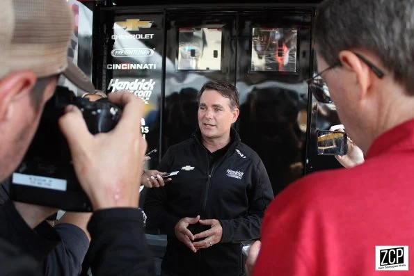 Jeff Gordon in dark Hendrick Motorsports team clothing, being photographed by reporters at a racetrack.