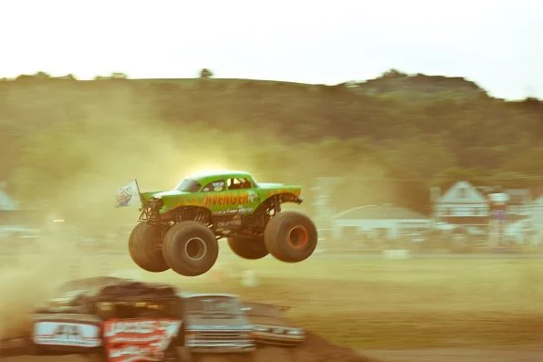 Jim Koehler's monster truck Avenger airborne over a line of crushed cars in a cloud of dust at an outdoor dirt track.