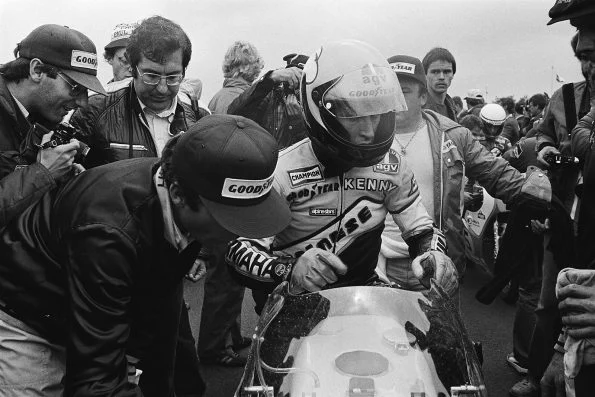 Kenny Roberts in helmet and leathers marked Kenny, leaning over his Yamaha in the paddock surrounded by photographers and crew in Goodyear caps.