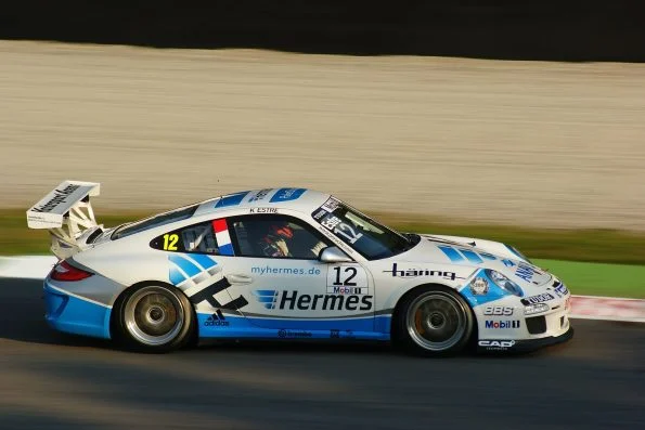 Kévin Estre driving a white and blue Hermes-liveried Porsche 911 GT3 Cup car, number 12, through the Ascari chicane at Monza.