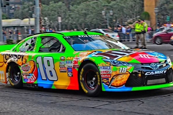 Kyle Busch's number 18 Toyota Camry in bright Skittles colours driving the NASCAR Victory Lap along the Las Vegas Strip, with a police escort behind.