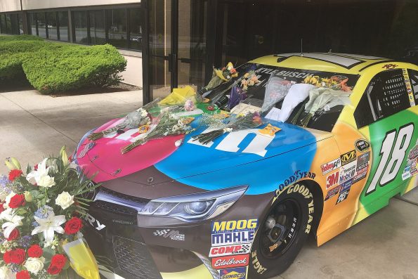 Kyle Busch's M&M's number 18 Toyota Camry on display outside a building, with bouquets of flowers and handwritten notes laid across its bonnet and roof.