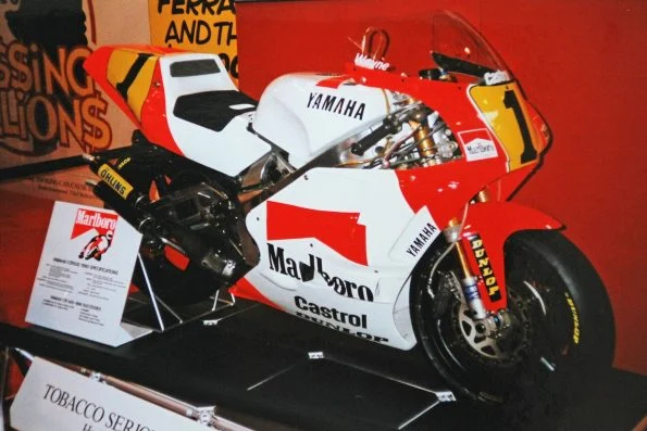 Wayne Rainey’s 1990 white and red Marlboro Yamaha YZR500, featuring the yellow number 1, displayed on a stand beside its specification card.