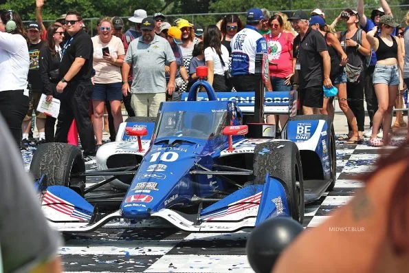 Álex Palou's blue American Legion Chip Ganassi Dallara-Honda, car 10, standing in victory lane on the chequered ground at Road America in 2023, confetti scattered across it and spectators crowding the barrier.