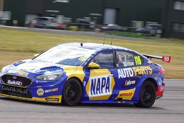 The blue and yellow number 116 NAPA Racing UK Ford Focus of Ash Sutton at Thruxton in 2026, pit garages behind.