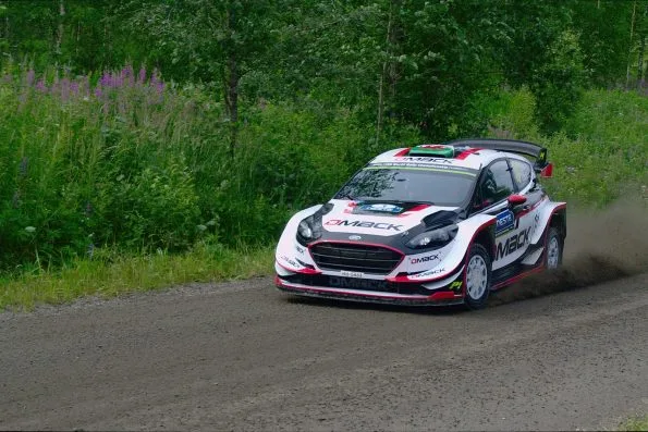 Elfyn Evans's white and red DMACK-liveried Ford Fiesta WRC turning in on the gravel of the Saalahti stage at Rally Finland in 2017, fireweed on the bank behind.