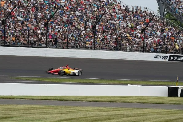 Josef Newgarden's red and yellow Shell Team Penske car, number 2, running through turn four at the Indianapolis Motor Speedway during the 2023 Indianapolis 500, a packed grandstand behind.