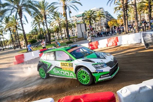 Kalle Rovanperä's green and white Škoda Fabia R5 evo, car 21, sliding through a palm-lined seafront section at Rally de España in 2019, his and Jonne Halttunen's names on the rear side window.
