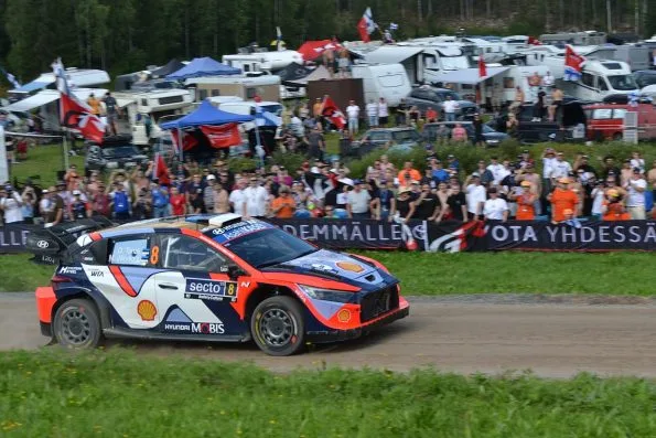 Ott Tänak's Hyundai i20 N Rally1, car 8, on the gravel of the Ouninpohja stage at Rally Finland in 2025, passing a packed spectator bank of camper vans and flags, his and Martin Järveoja's names on the side window.