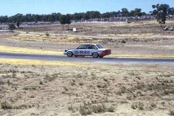 Peter Brock driving the white, red and blue number 05 Mobil Holden Dealer Team Commodore on track at Wanneroo Raceway in 1985, with spectators lining the far bank