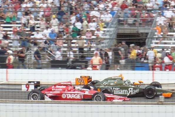 Scott Dixon taking his victory lap in the number 9 Target car after winning the 2008 Indianapolis 500, a packed grandstand behind him and the green number 99 car alongside.
