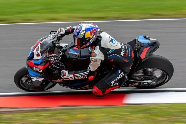 Razgatlıoğlu leaned hard over with his elbow down on the number 54 black, blue and red ROKiT BMW M1000RR at Donington Park in July 2024, the red kerb blurred beneath him